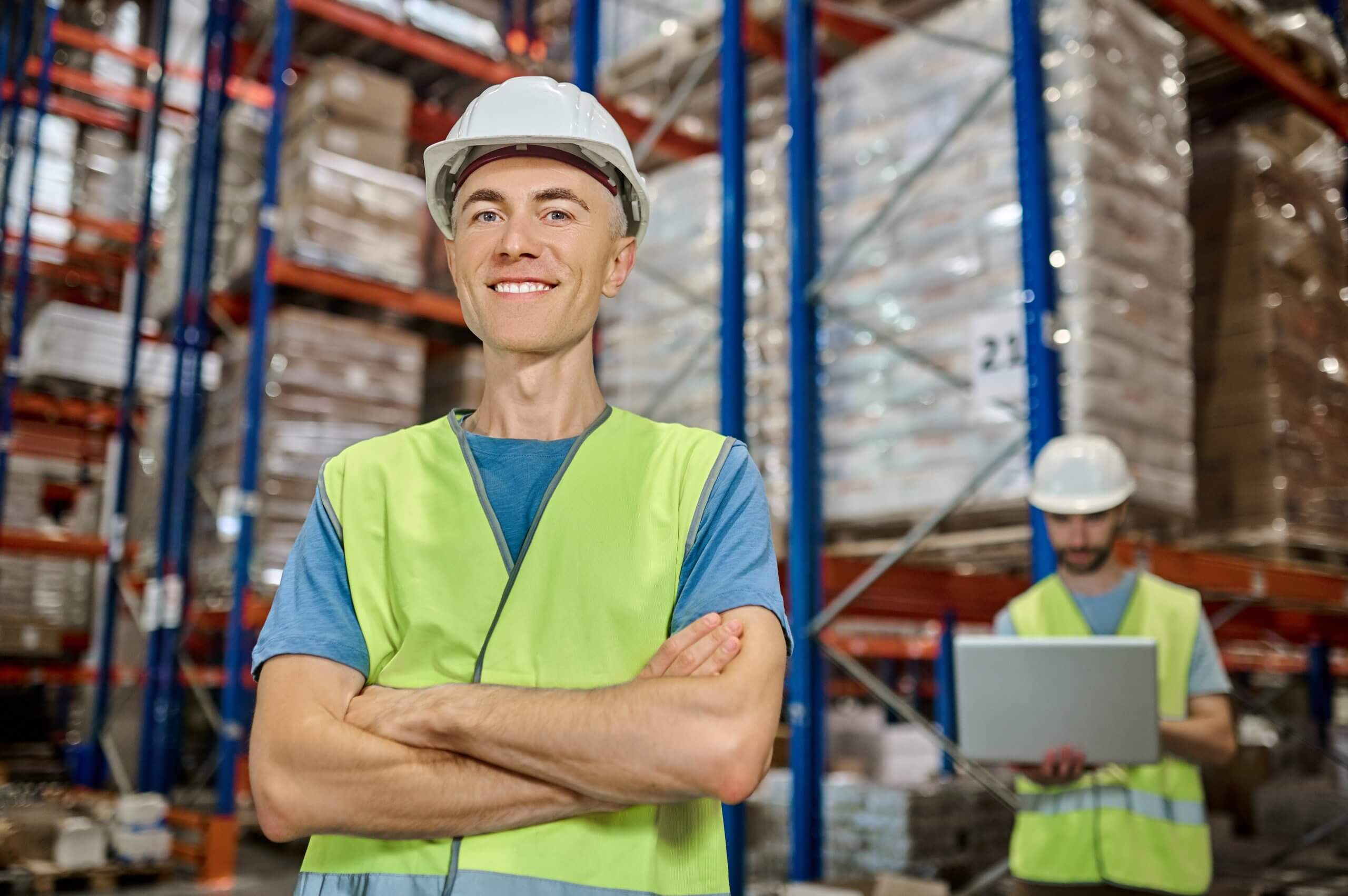 All perfectly. Young adult smiling man in protective helmet and workwear standing with folded hands looking at camera and colleague working on laptop behind