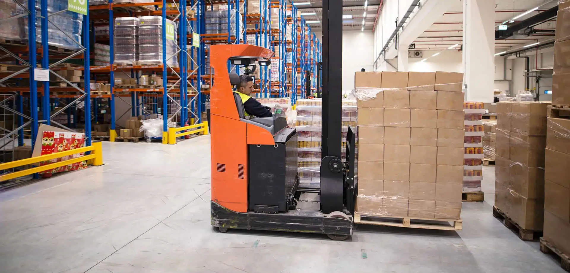 A worker operates a red and black electric reach truck in a clean warehouse, moving a pallet of boxes between tall shelving units stocked with goods.