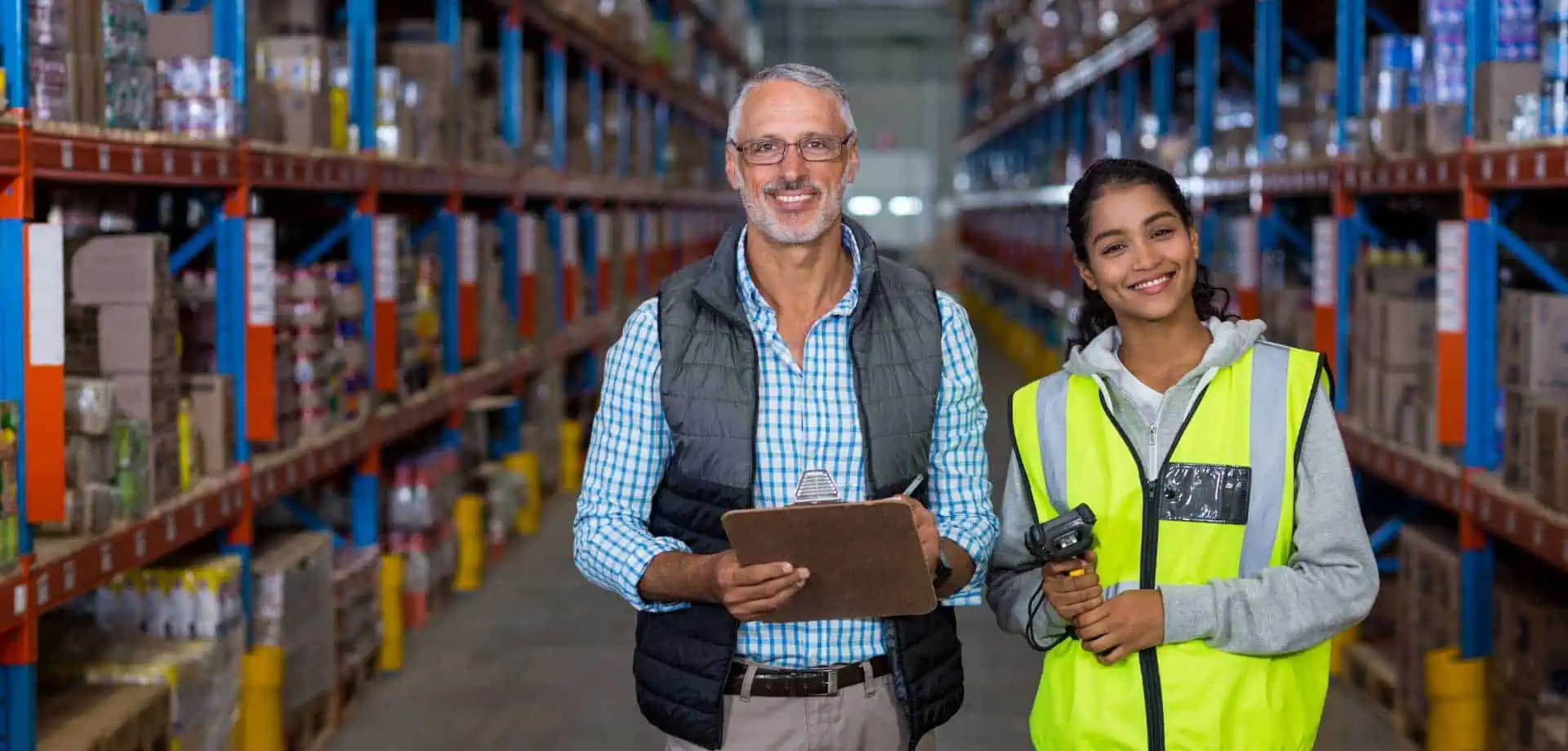 Two logistics workers stand in a warehouse aisle lined with stocked shelves. One holds a clipboard and wears a vest, while the other wears a high-visibility safety vest and holds a barcode scanner. Both are smiling, highlighting teamwork in inventory and supply chain operations.